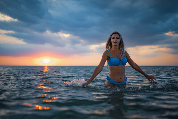 A girl with blond hair in a blue swimsuit splashes to the sides while sitting in an estuary on a sunset background - Foto, imagen