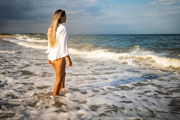 A slender girl in a gentle blue swimsuit and shirt walks along the sandy beach near the blue sea - Photo, image