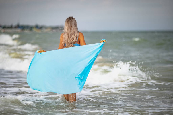 Mavi mayolu, sarı saçlı ve parlak şallı bir kız kumsalda yürüyor. - Fotoğraf, Görsel