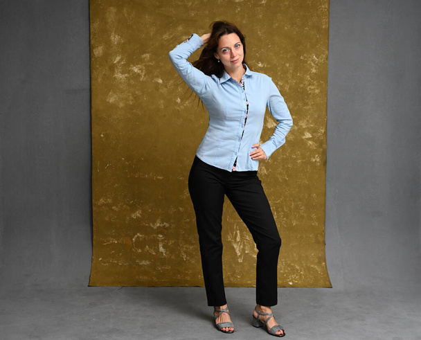 Full length photo of a happy brunette woman in a blue shirt and black trousers on a gray-golden background - Fotografie, Obrázek