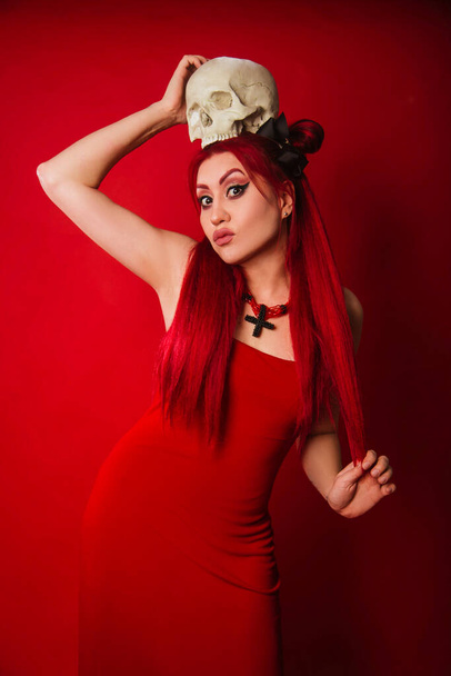 Informal Asian woman in a red evening dress with a skull in her hands. Modern witch with red hair. Halloween look - Photo, Image
