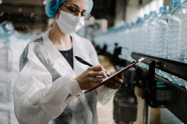 Pracownica z maską ochronną pracująca w fabryce zaopatrzenia medycznego i produkcji oraz sprawdzająca pojemniki z wodą destylowaną przed wysyłką. Kontrola jakości inspekcji.  - Zdjęcie, obraz
