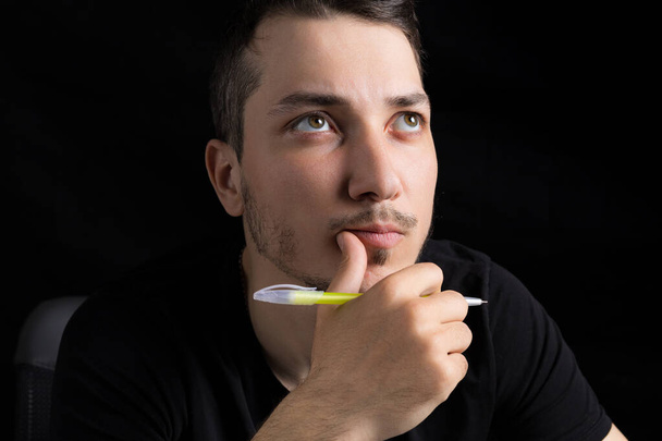 A young handsome man in black clothes with a pen looks up thoughtfully and dreamily, on a black background in a low key. Serious marketing and planning concept - Photo, Image