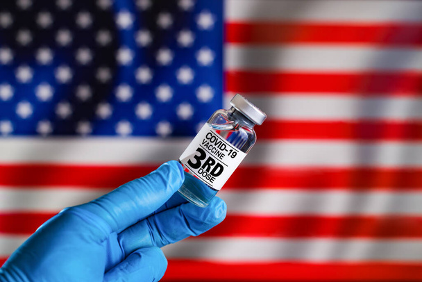 Trzecia dawka szczepionki Covid-19 do szczepienia przypominającego w chorobach populacji ryzyka w Stanach Zjednoczonych. Lekarz z fiolką z trzecią dawką szczepionki przeciw kovidowi-19 lub koronawirusowi przed flagą USA - Zdjęcie, obraz