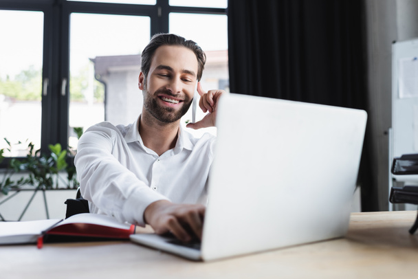 happy businessman using blurred laptop while talking on smartphone in office - Fotó, kép