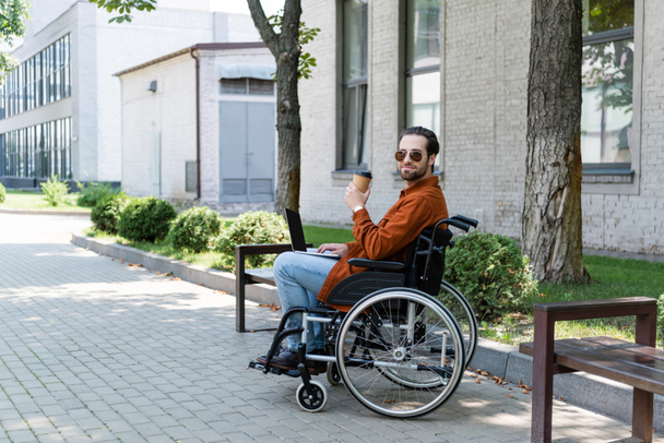 młody niepełnosprawny mężczyzna siedzi na wózku inwalidzkim z laptopem i na wynos drink na świeżym powietrzu - Zdjęcie, obraz