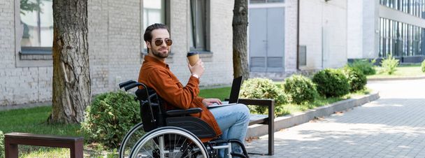 disabled man in sunglasses holding coffee to go while sitting in wheelchair with laptop, banner - Φωτογραφία, εικόνα