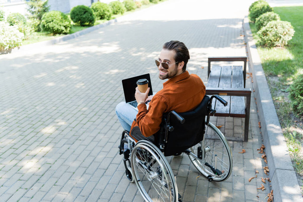 disabled man smiling at camera while sitting in wheelchair with paper cup and laptop - Φωτογραφία, εικόνα