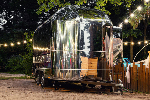 Scenic evening view of vintage futuristic silver metal camper van trailer parked near wooden fence at countryside farm or ranch against light garland forest tree. Retro style fodtruck caravan vehicle - Foto, Bild