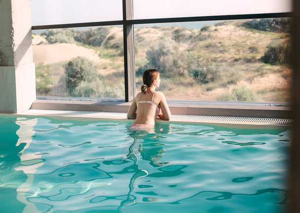 Chica joven nada en la piscina de un centro de spa de lujo junto a las grandes ventanas con vista. Spa, concepto de vacaciones - Foto, Imagen