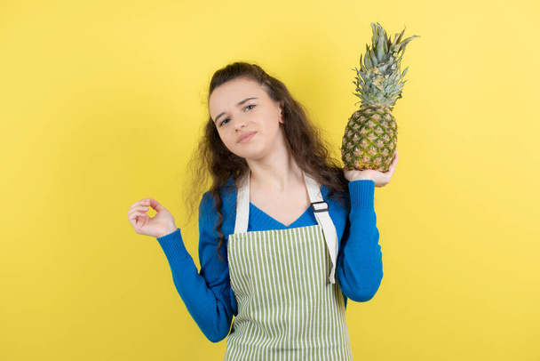 Photo d'un jeune adolescent bouclé dans un tablier tenant de l'ananas mûr au-dessus d'un mur jaune. Photo de haute qualité - Photo, image