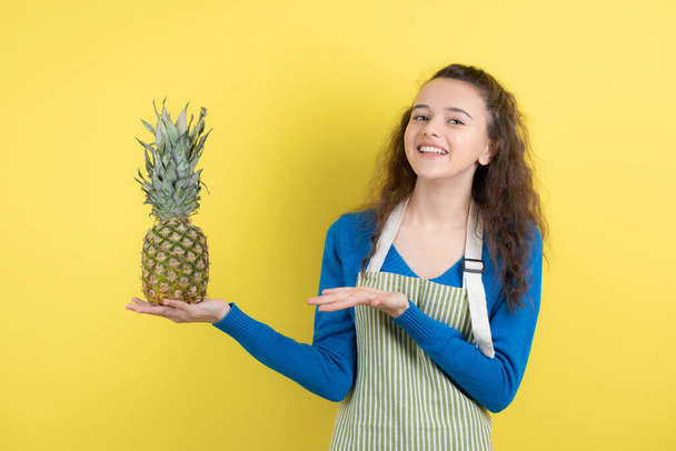 Jovem adolescente fêmea em avental segurando abacaxi maduro sobre a parede amarela. Foto de alta qualidade - Foto, Imagem