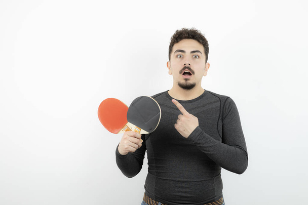 Foto pohledného mladého sportovce ukazujícího nahoru a držícího rakety pro stolní tenis. Kvalitní fotografie - Fotografie, Obrázek