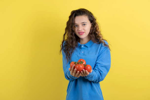 黄色の背景に立って赤いトマトと青のスウェットシャツの若い10代の女の子。高品質の写真 - 写真・画像