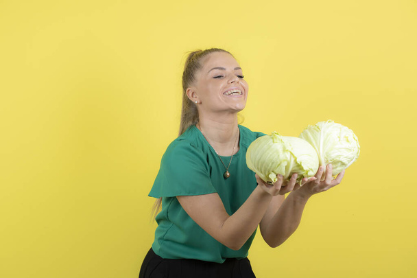 Image of young housewife holding heavy fresh cabbages over yellow wall. High quality photo - Fotoğraf, Görsel