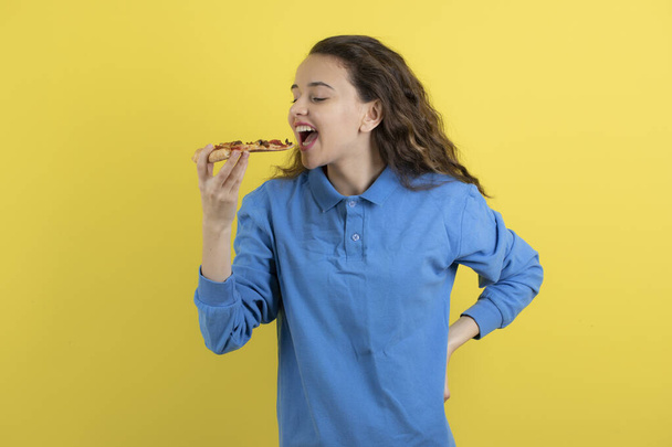 Photo of beautiful young girl eating piece of pizza over yellow wall. High quality photo - Photo, Image