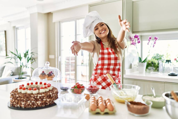 Красивая юная брюнетка-кондитер-повар готовит пирожные на кухне, глядя на камеру, улыбающуюся с распростертыми объятиями для объятий. веселое выражение, охватывающее счастье.  - Фото, изображение
