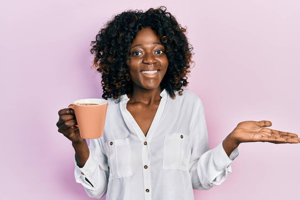 Joven mujer afroamericana sosteniendo café celebrando logro con sonrisa feliz y expresión ganadora con la mano levantada  - Foto, imagen