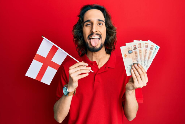 Jonge Latijns-Amerikaanse man met Engelse vlag en ponden bankbiljetten steken tong uit gelukkig met grappige uitdrukking.  - Foto, afbeelding