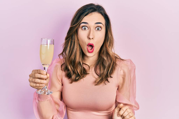 Chica hispana joven bebiendo una copa de champán espumoso asustado y sorprendido con la boca abierta para sorpresa, cara de incredulidad  - Foto, imagen