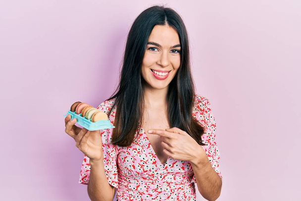 jong brunette vrouw holding heerlijk macarons gebak glimlachen gelukkig wijzend met de hand en vinger  - Foto, afbeelding