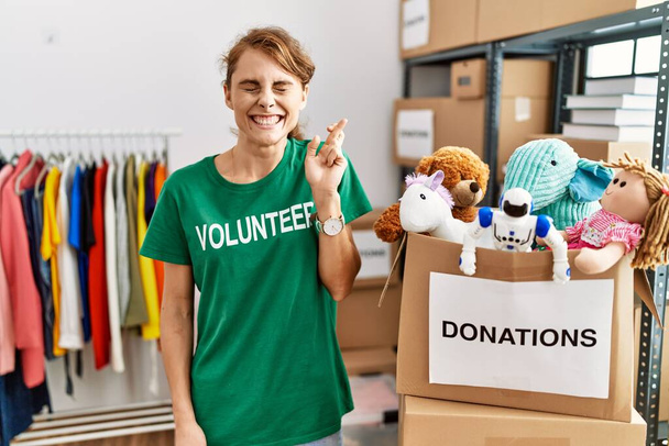 Красивая кавказка в волонтерской футболке на пожертвованиях стоит, жестом скрестив пальцы, улыбаясь с надеждой и закрыв глаза. удача и суеверие.  - Фото, изображение