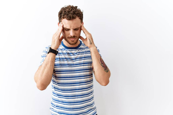 Knappe jongeman die over een geïsoleerde achtergrond staat met hand op hoofd, hoofdpijn door stress. lijden aan migraine.  - Foto, afbeelding