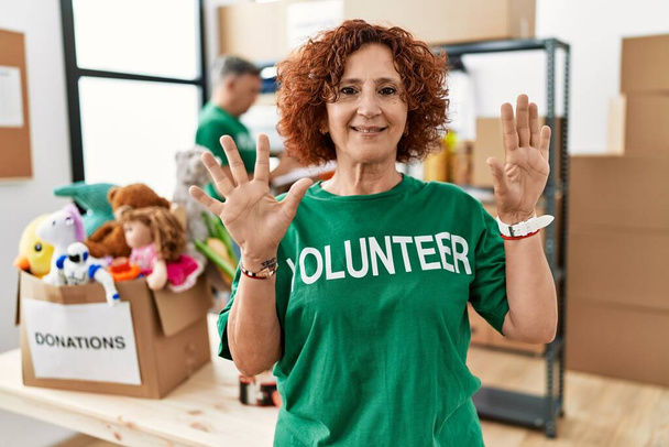 Femme d'âge moyen portant un t-shirt volontaire sur le stand de dons montrant et pointant vers le haut avec les doigts numéro dix tout en souriant confiant et heureux.  - Photo, image