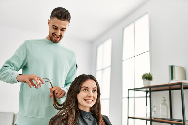 Jeune styliste hispanique homme coupe les cheveux de la femme en utilisant des ciseaux au centre de beauté. - Photo, image