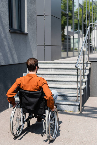 widok z tyłu młody niepełnosprawny mężczyzna na wózku inwalidzkim w pobliżu budynku ze schodami - Zdjęcie, obraz