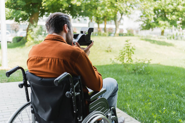 back view of handicapped man in wheelchair taking photo on vintage camera outdoors - Фото, изображение