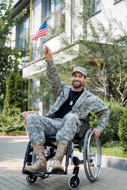 soldado alegre con pequeña bandera de EE.UU. en la mano levantada mirando a la cámara al aire libre - Foto, Imagen
