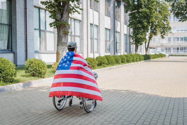 Rückansicht eines behinderten Soldaten, der in eine US-Flagge gehüllt im Rollstuhl auf einer städtischen Straße sitzt - Foto, Bild