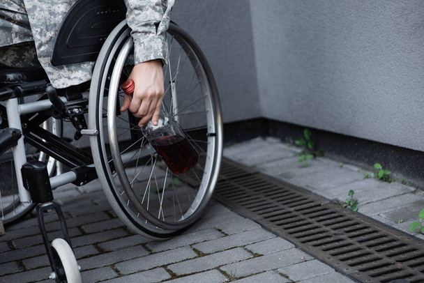 partial view of handicapped military man with bottle of whiskey sitting in wheelchair outdoors - Foto, Bild