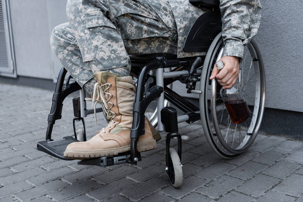vista recortada del soldado discapacitado en silla de ruedas sosteniendo botella de whisky al aire libre - Foto, Imagen