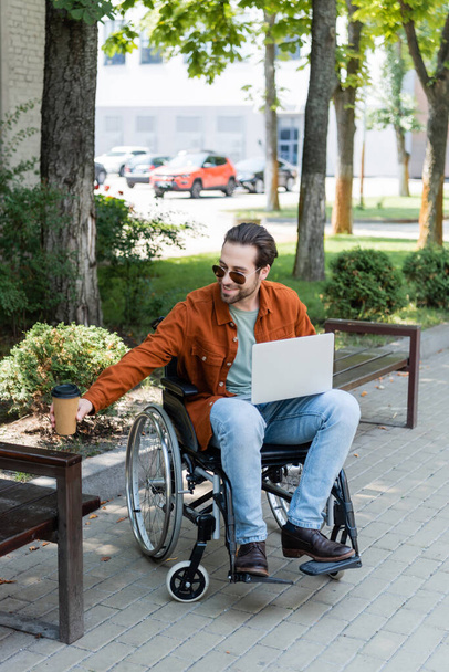 sonriente hombre discapacitado sosteniendo café para ir sentado en silla de ruedas con el ordenador portátil - Foto, Imagen