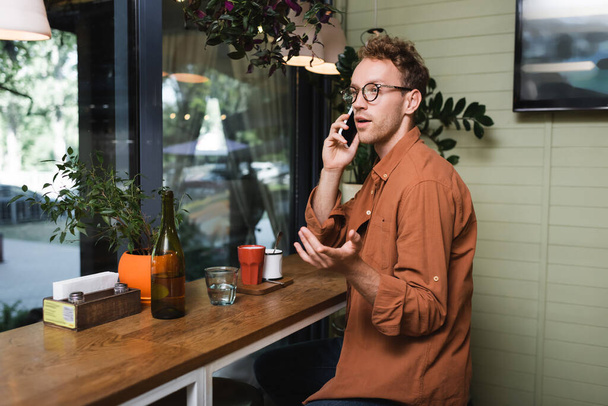 joven en gafas haciendo gestos mientras habla en el teléfono inteligente en la cafetería  - Foto, Imagen