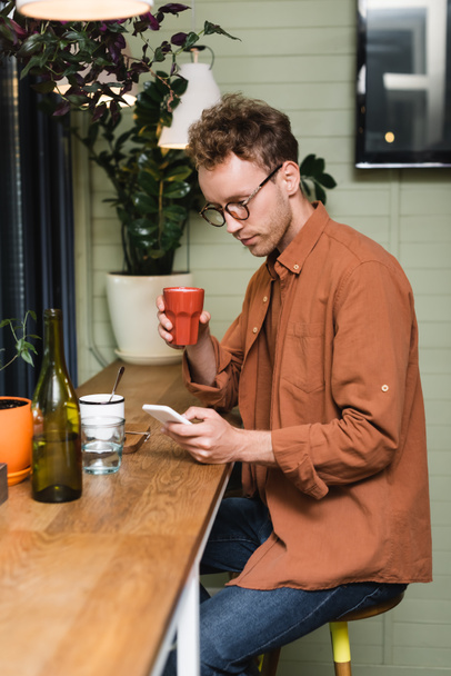 Młody człowiek w okularach smsuje na smartfonie w kawiarni  - Zdjęcie, obraz