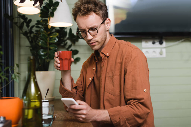 young man in glasses holding cup and texting on smartphone in cafe  - Fotografie, Obrázek