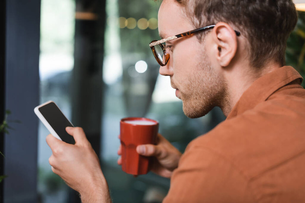 young man in glasses holding cup of coffee and texting on smartphone in cafe  - Foto, imagen