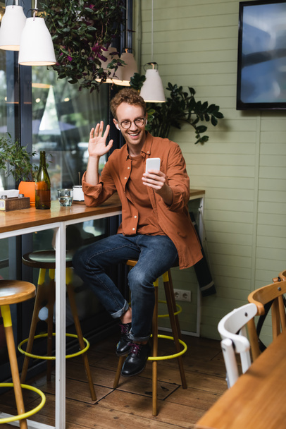 šťastný mladý muž v brýlích a bezdrátových sluchátkách mává rukou během videohovoru v kavárně - Fotografie, Obrázek