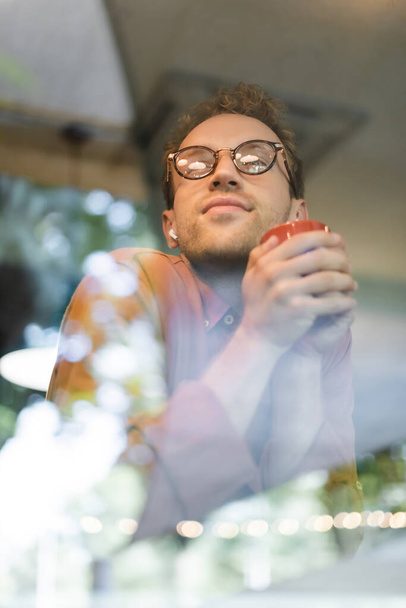 άποψη χαμηλή γωνία του νεαρού άνδρα σε γυαλιά και ακουστικά κρατώντας φλιτζάνι καφέ πίσω από θολή παράθυρο του καφέ - Φωτογραφία, εικόνα