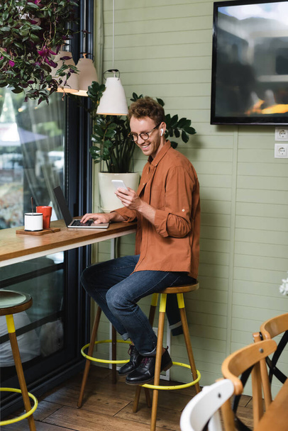 happy young freelancer in glasses using smartphone near laptop in cafe - Foto, afbeelding