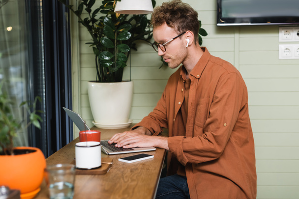 young freelancer in glasses typing on laptop near smartphone in cafe - Photo, Image