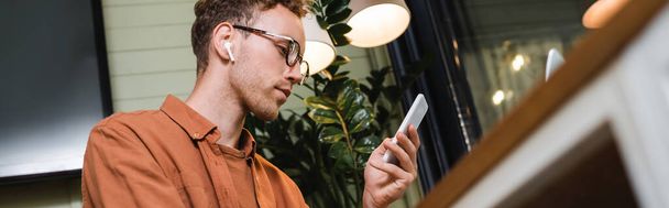 niski kąt widzenia młodego człowieka w okularach i słuchawkach bezprzewodowych za pomocą smartfona w kawiarni, baner - Zdjęcie, obraz
