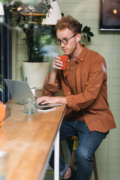 νεαρός ελεύθερος επαγγελματίας σε ποτήρια κρατώντας κύπελλο και πληκτρολογώντας σε φορητό υπολογιστή κοντά στο smartphone στο καφέ - Φωτογραφία, εικόνα
