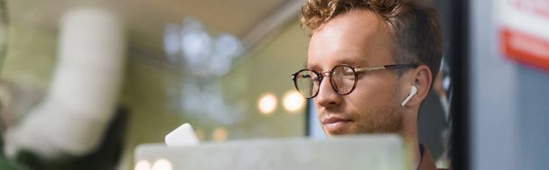 young man in glasses and earphones near laptop behind blurred window of cafe, banner - Фото, изображение
