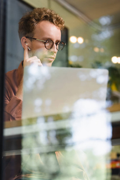 young man in glasses adjusting earphones near laptop behind blurred window of cafe - Фото, изображение