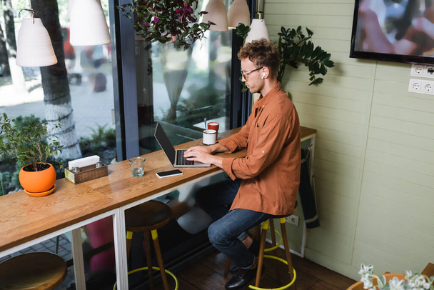 фрилансер в наушниках с помощью ноутбука рядом с сотовым телефоном и напитки на столе в кафе  - Фото, изображение