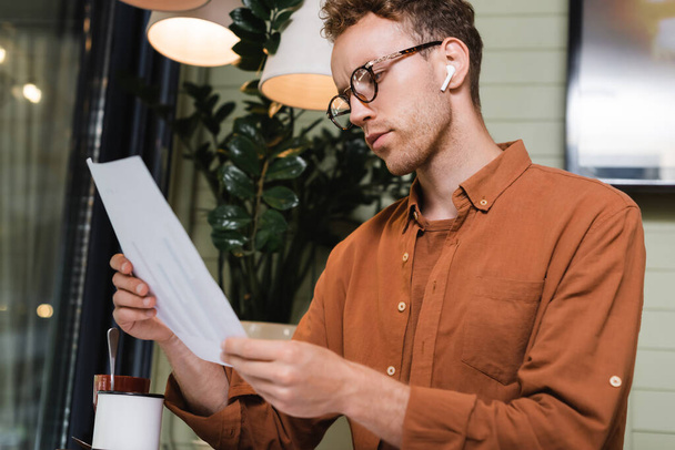 young man in glasses and earphones looking at paper with graphs in cafe  - Photo, Image
